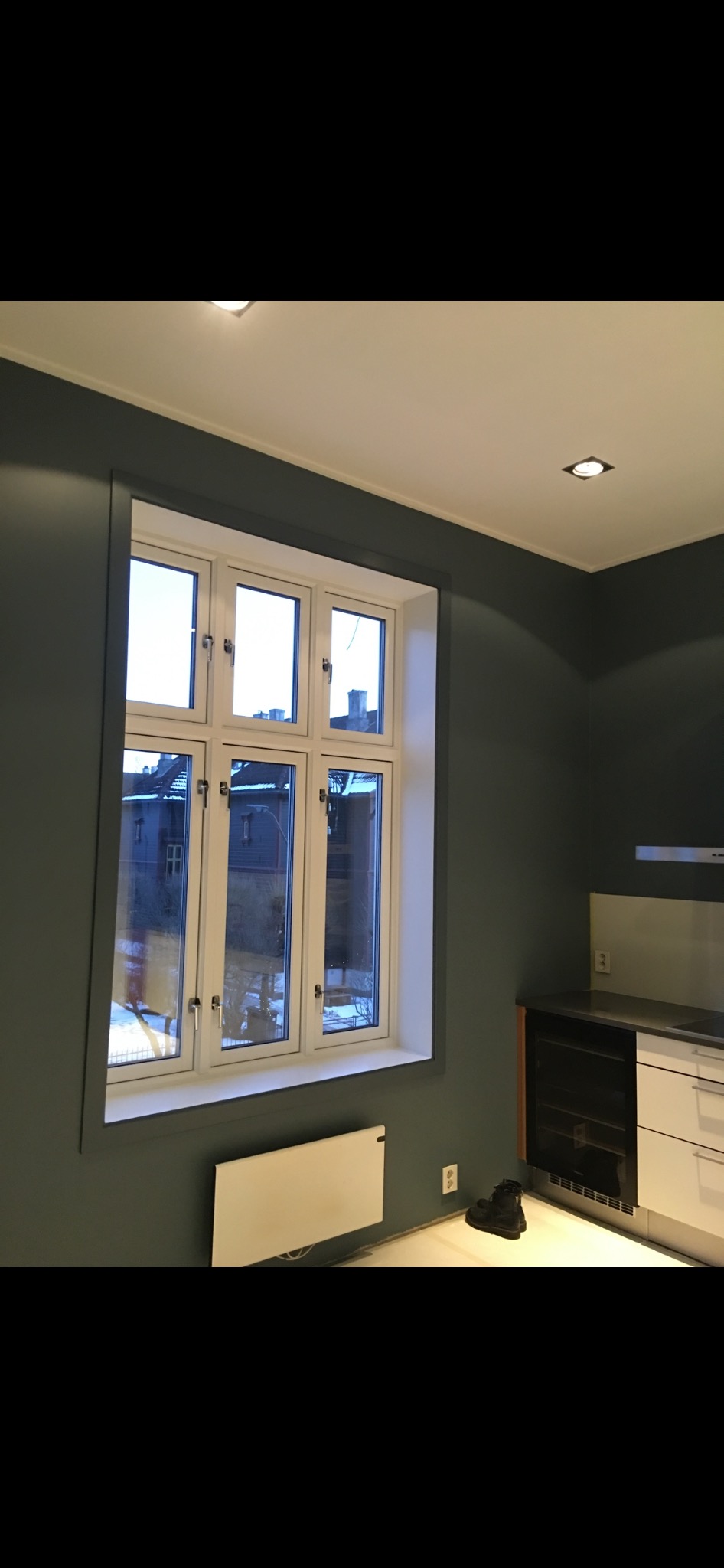 Pomieszczenie po malowaniu ścian na ciemnozielony kolor, widoczne okno z białymi ramami, grzejnik pod oknem i fragment zabudowy kuchennej z jasnymi frontami.