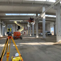 Pomiar geodezyjny wewnątrz hali w trakcie budowy, teodolit na statywie z żółtymi nogami na pierwszym planie, w tle podnośnik z pracownikami przy konstrukcji dachu, widoczny samochód dostawczy.