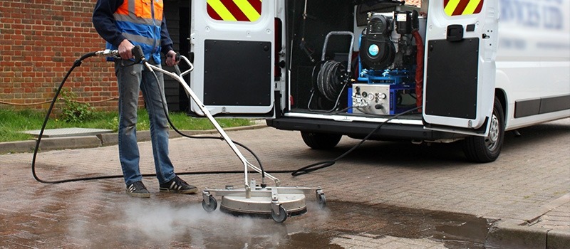 Pracownik w odblaskowej kamizelce czyści kostkę brukową za pomocą profesjonalnej myjki ciśnieniowej z dyszą rotacyjną; w tle otwarty samochód dostawczy z zamontowanym agregatem myjącym.