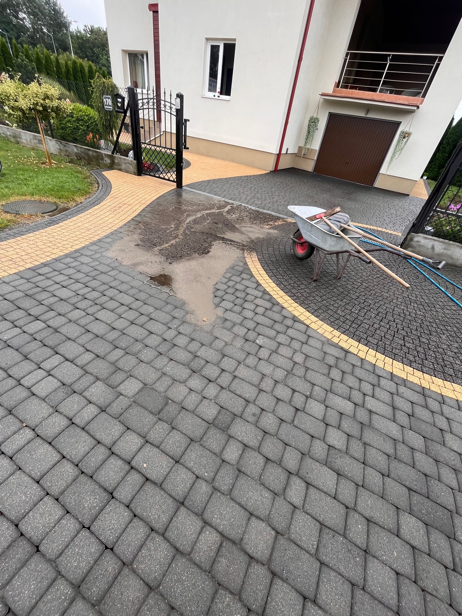 Częściowo umyta, mokra kostka brukowa w kolorze szarym i żółtym przed domem jednorodzinnym. W tle brama i wózek z narzędziami.