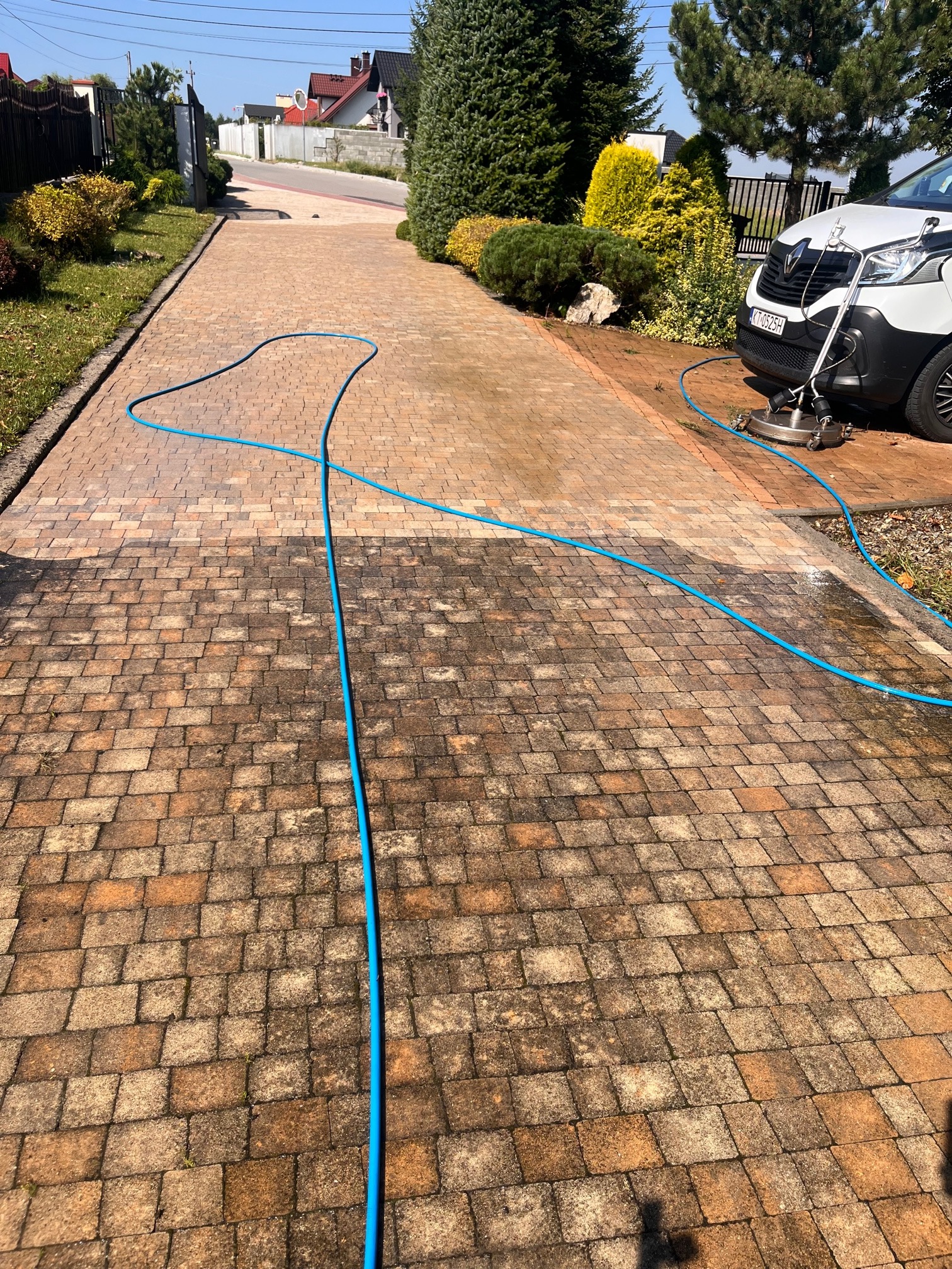 Czyszczenie kostki brukowej z użyciem myjki ciśnieniowej; widoczny fragment posesji z ogrodem i samochodem dostawczym firmy.
