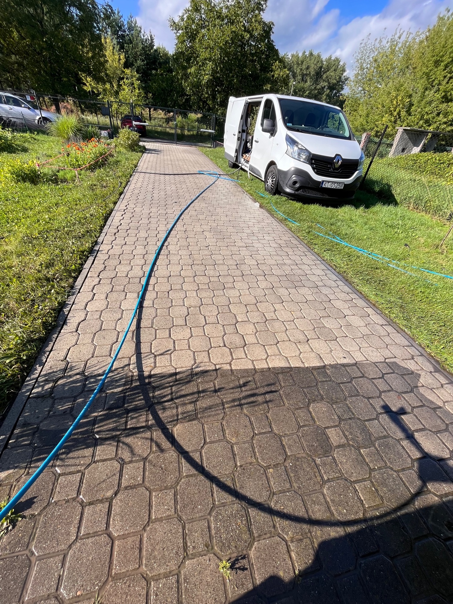 Częściowo umyta kostka brukowa z wyraźnym kontrastem czystej i brudnej powierzchni, niebieski wąż ciśnieniowy ciągnący się wzdłuż ścieżki, biały samochód dostawczy firmy parkujący na trawie w tle.