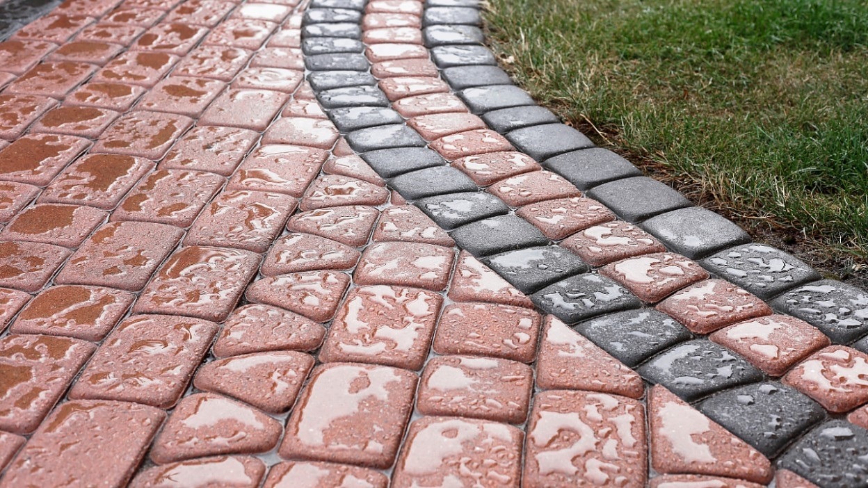 Mokra kostka brukowa w odcieniach czerwieni i szarości, ułożona w łuk przy trawniku, widoczne krople wody na powierzchni.