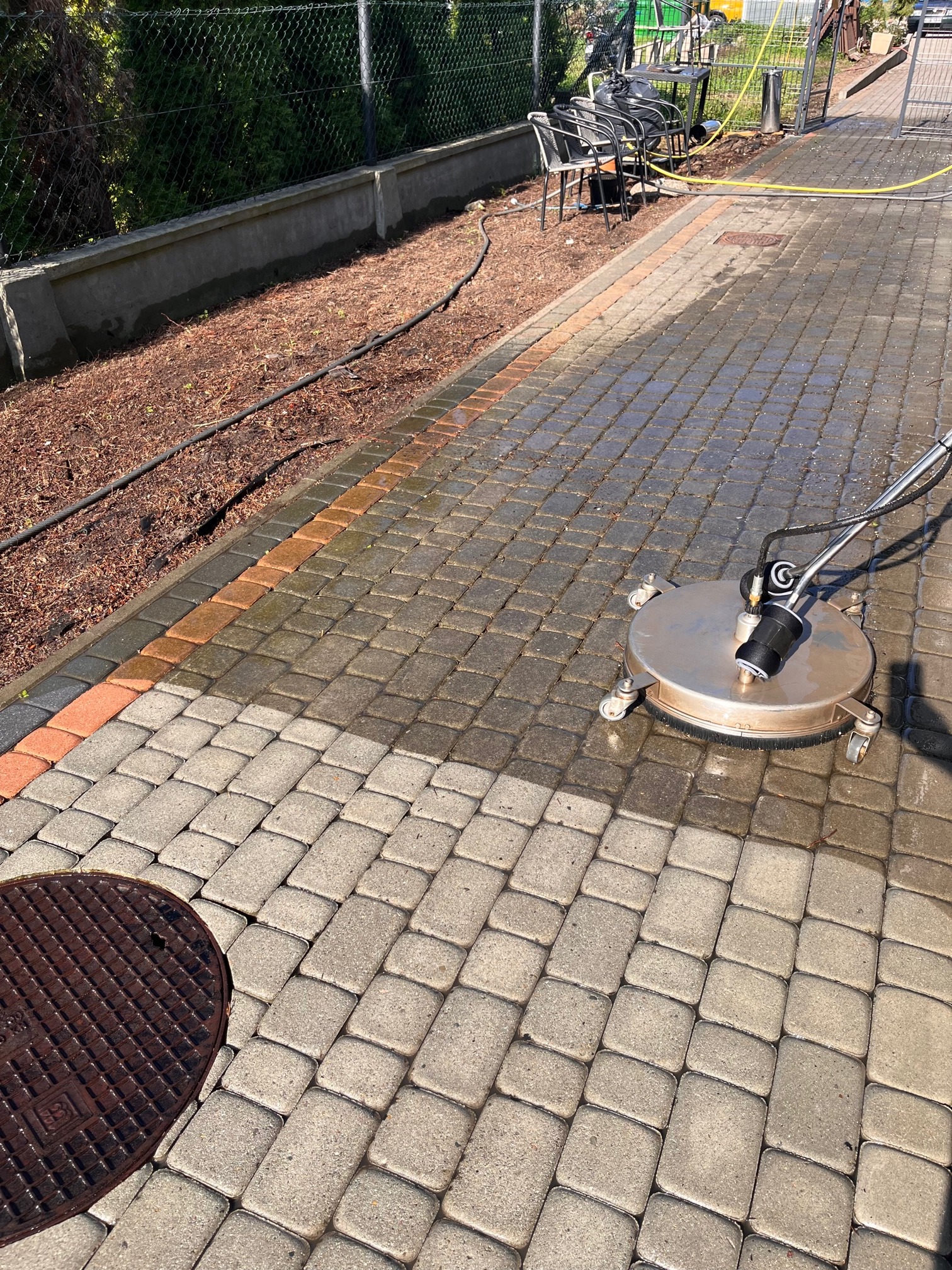 Czyszczenie kostki brukowej przy użyciu okrągłej myjki ciśnieniowej. Widoczna różnica w kolorze pomiędzy umytą a brudną powierzchnią kostki. W tle ogrodzenie i elementy wyposażenia ogrodu.