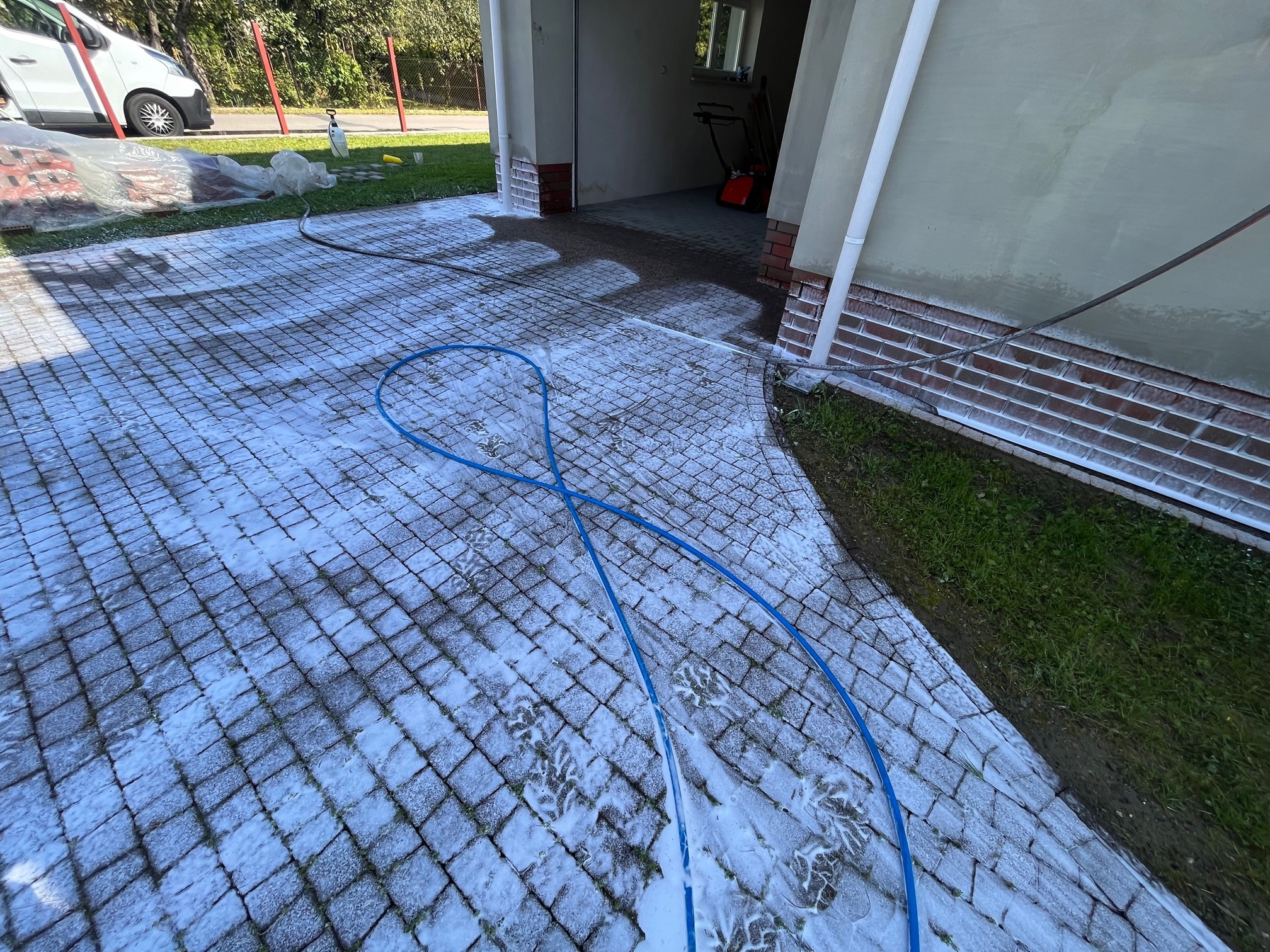 Prywatna posesja: Czyszczenie kostki brukowej pokrytej pianą z widocznymi śladami butów, niebieski wąż ciśnieniowy ułożony na bruku, zaparkowany biały samochód dostawczy w tle.