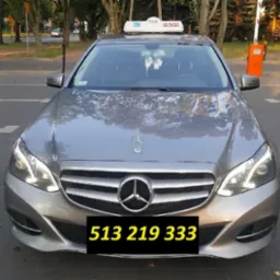 Srebrny Mercedes-Benz z lampką Taxi na dachu, zaparkowany przy ulicy, widok z przodu.