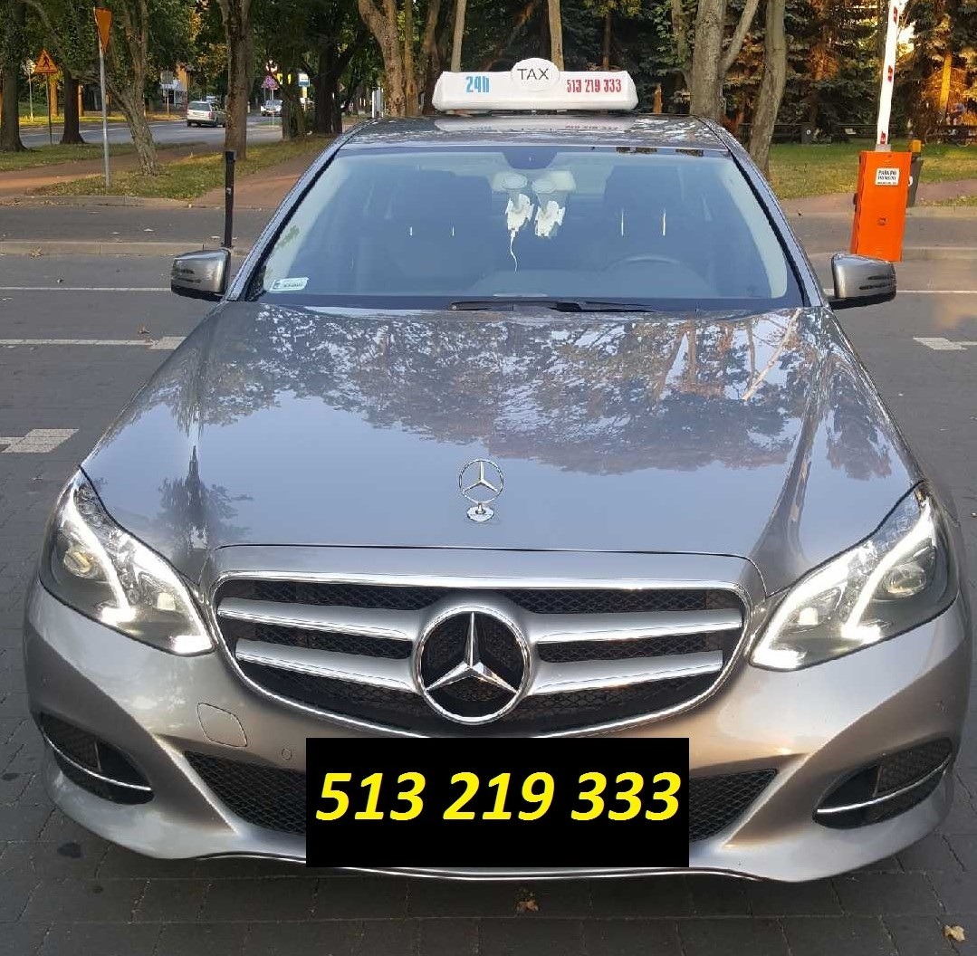 Srebrny Mercedes-Benz z lampką Taxi na dachu, zaparkowany przy ulicy, widok z przodu.