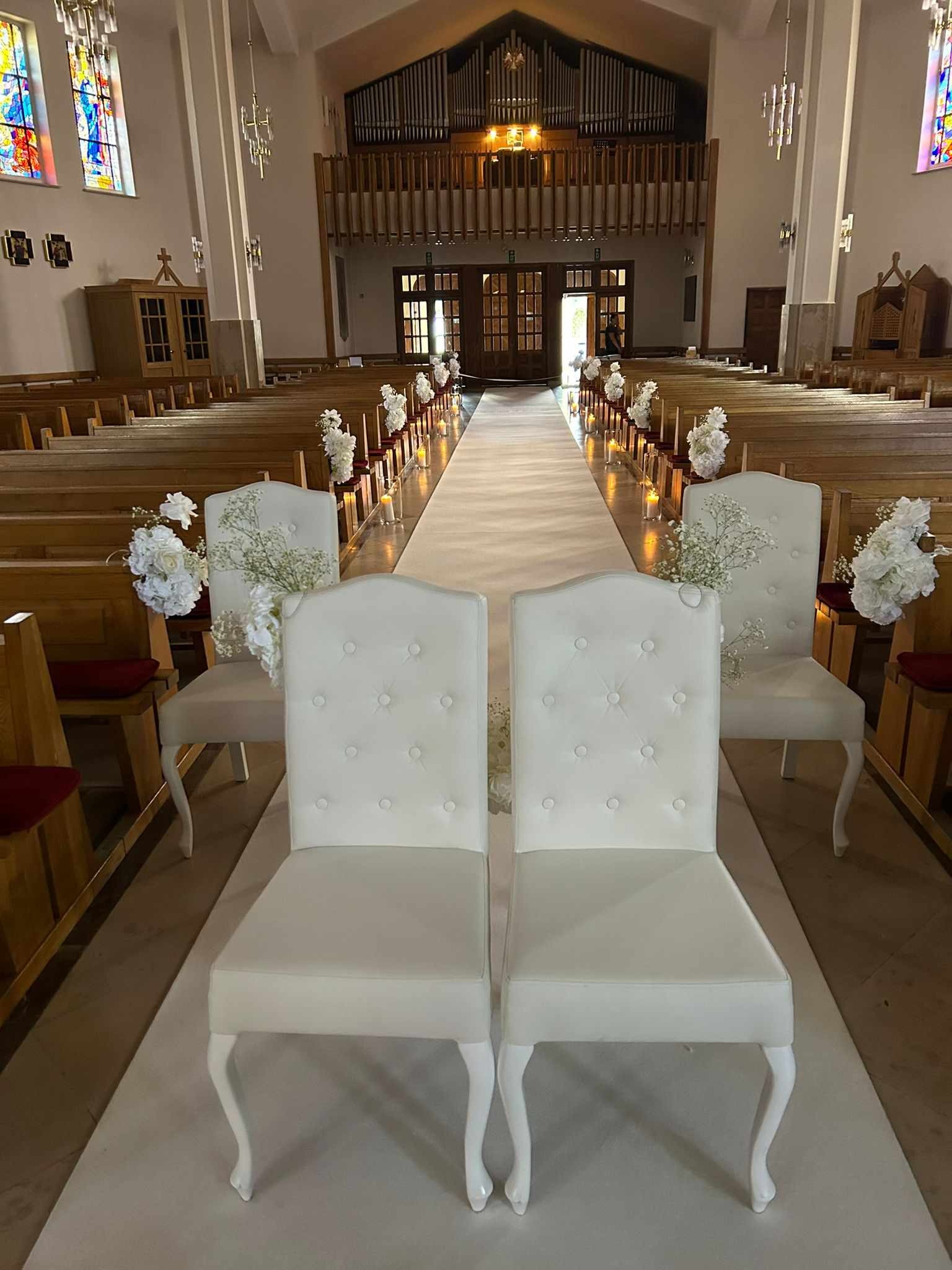 Dekoracja kościoła na uroczystość ślubną: biały dywan, krzesła z białym obiciem i kwiatami, oświetlenie świecami.