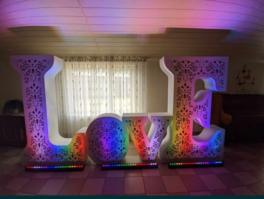 Duże, podświetlane litery LOVE z ażurowym wzorem, ustawione na podłodze w pomieszczeniu z zasłonami w tle, tworzące dekorację eventową.