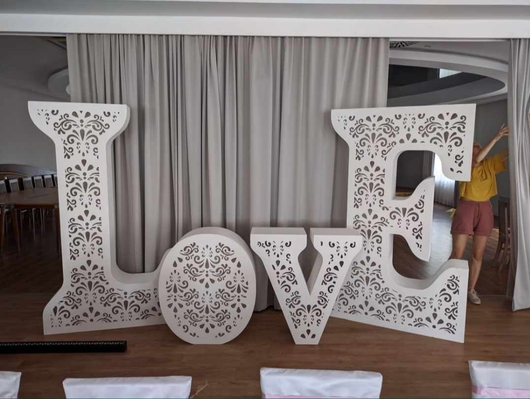 Dekoracyjne, ażurowe, białe litery LOVE ustawione na drewnianej podłodze, z zasłonami w tle i osobą w żółtej koszulce w tle, przygotowane na imprezę.