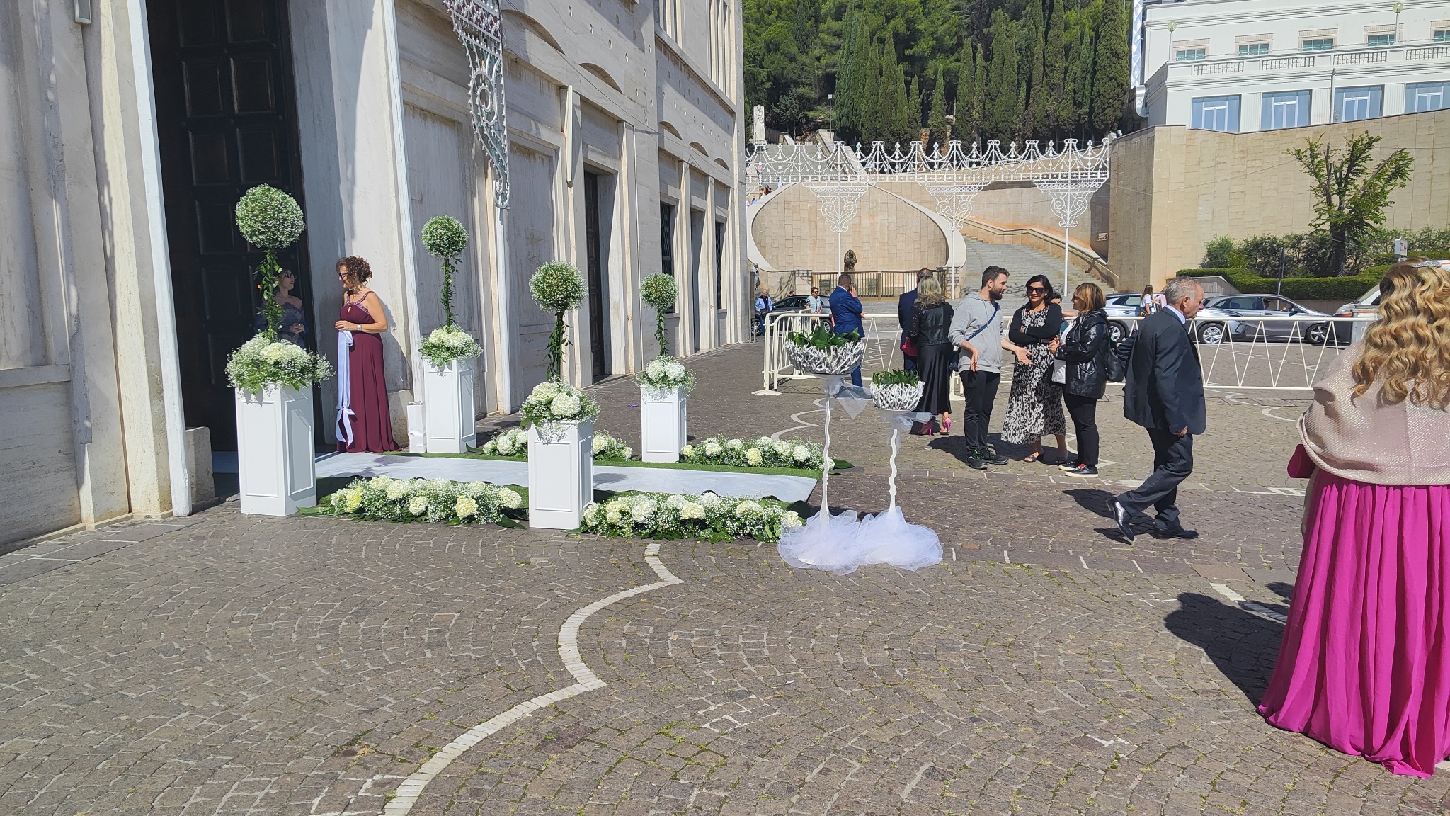 Dekoracja kwiatowa przed wejściem do kościoła, przygotowania do uroczystości ślubnej, goście weselni w tle.