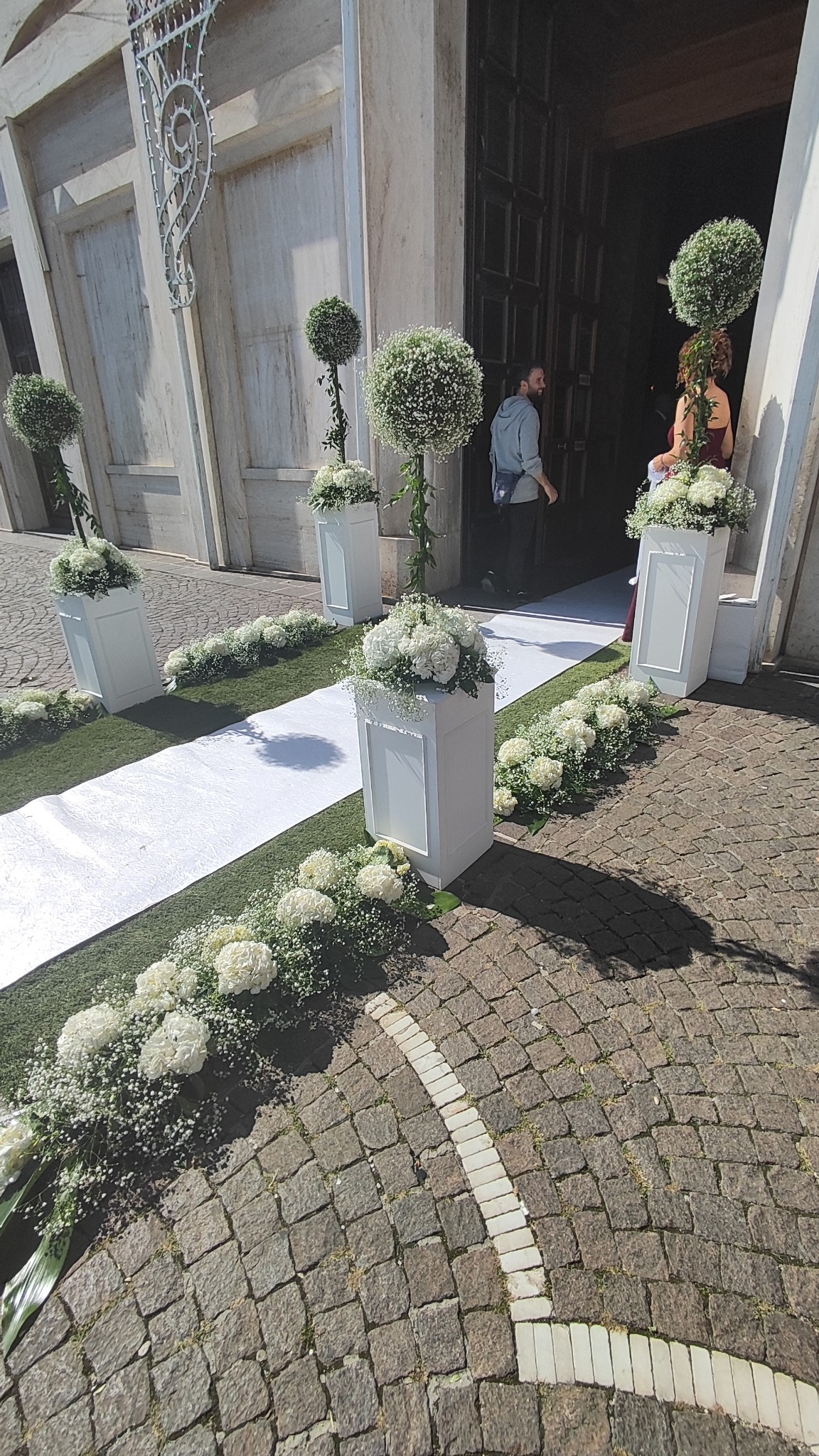 Dekoracja ślubna przed wejściem do kościoła: biały dywan, kompozycje kwiatowe w białych donicach, topiary z zielonych kul.