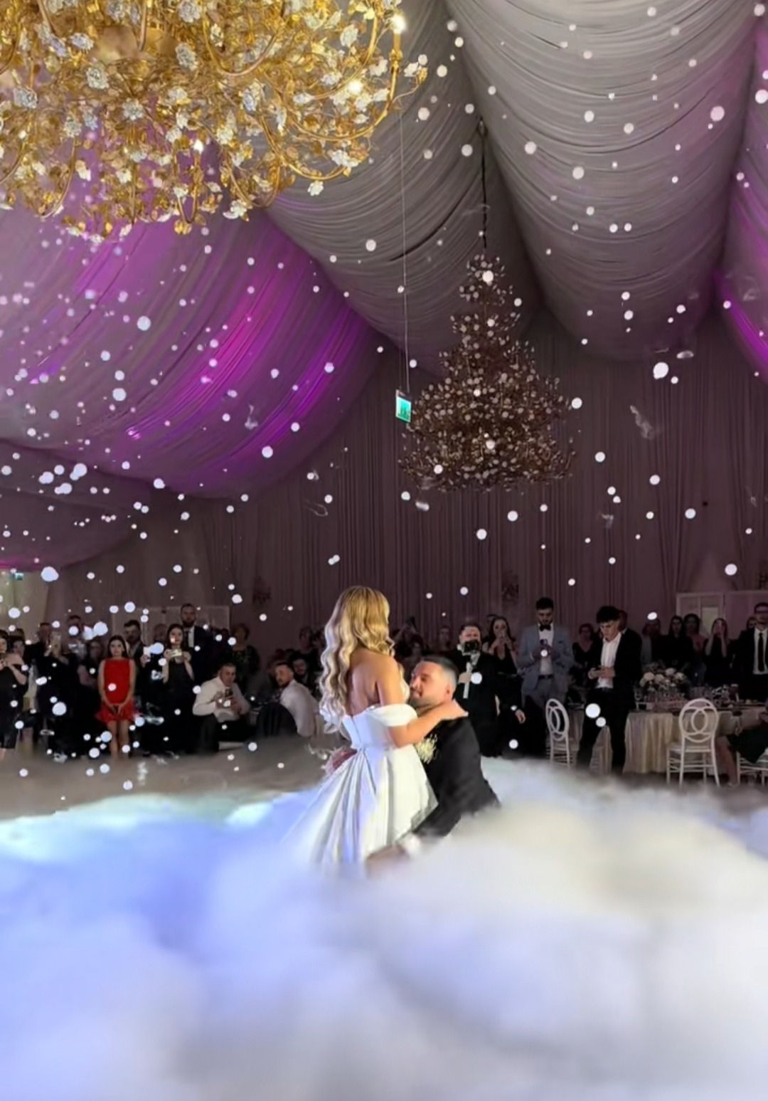 Para młoda tańczy pierwszy taniec w chmurach na sali weselnej z dekoracjami i gośćmi w tle. Efekt specjalny z ciężkiego dymu i konfetti.