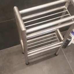 Stojak łazienkowy ze stali nierdzewnej z dwoma półkami, umieszczony na szarej podłodze przy ciemnej ścianie, widoczna stopa osoby w jeansach.