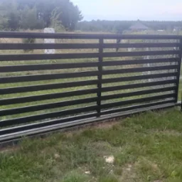 Czarna, przesuwna brama wjazdowa z poziomymi panelami, widoczna z zewnątrz posesji, z mechanizmem jezdnym i podporami, na tle trawnika i ogrodzenia z siatki.