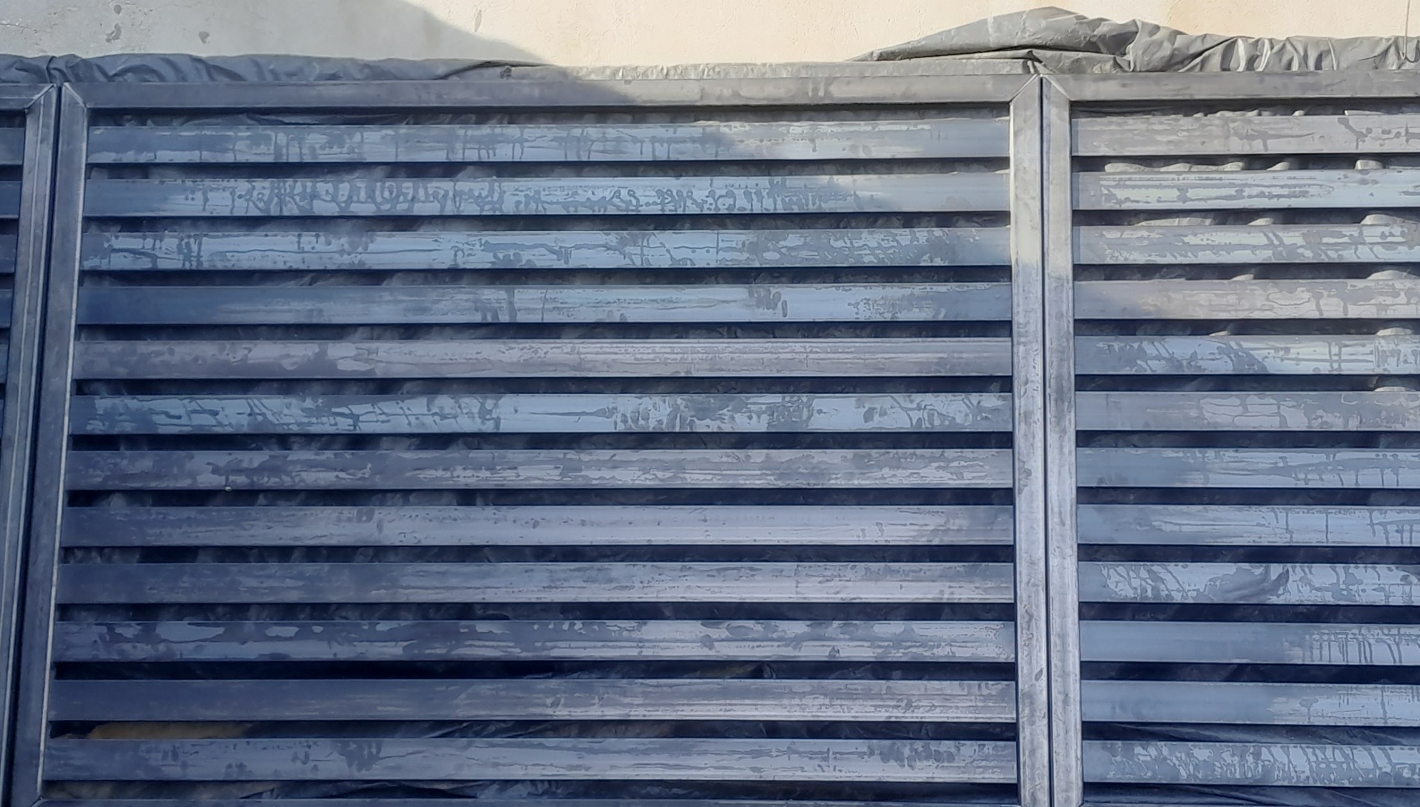 Metalowa konstrukcja z poziomych profili, prawdopodobnie fragment bramy lub ogrodzenia, w trakcie obróbki lub malowania, widoczne ślady po cieczach.