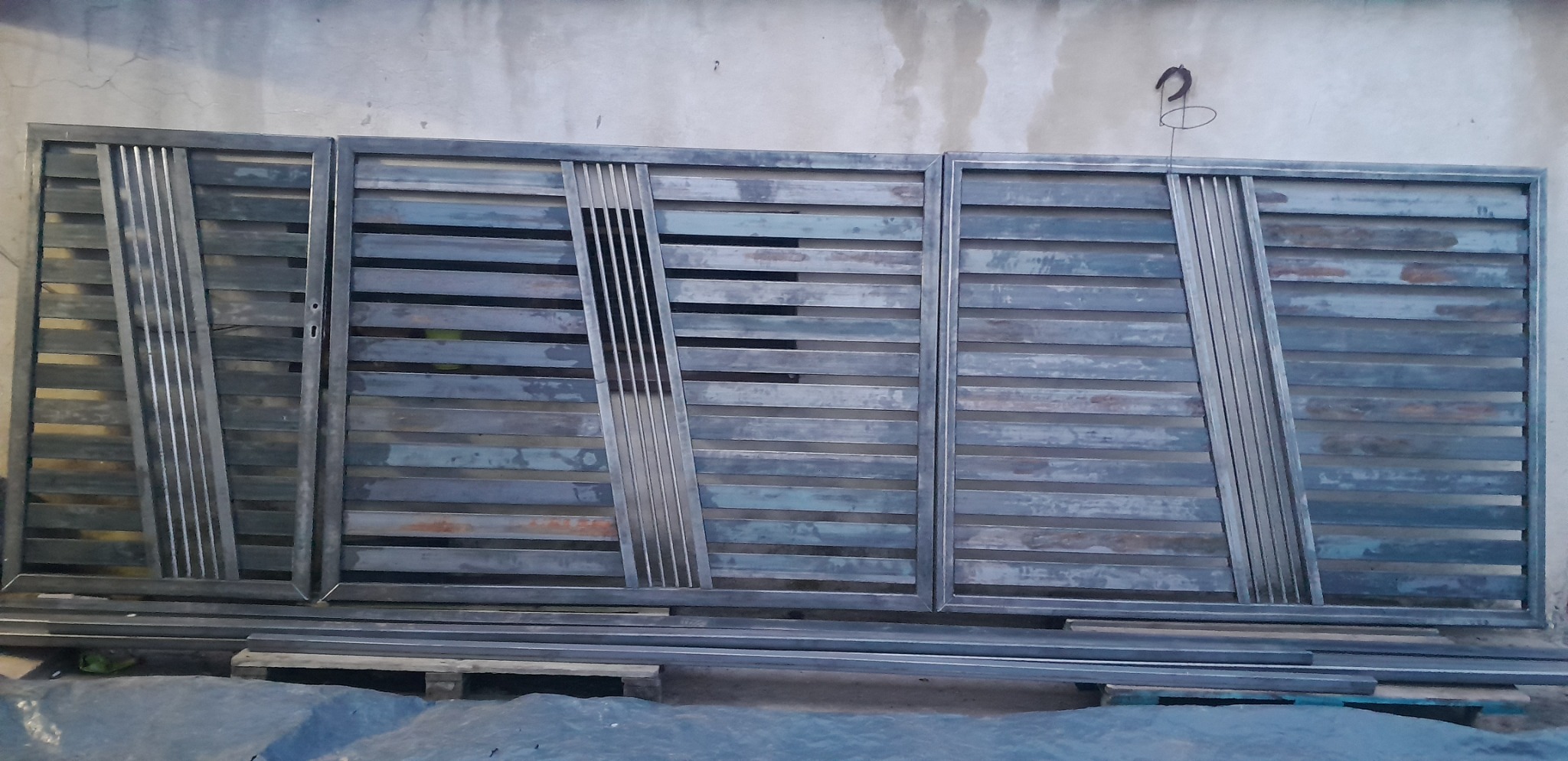 Surowa, metalowa brama wjazdowa z poziomymi prętami i ukośnymi wzmocnieniami, jeszcze nie zamontowana, leżąca na palecie pod ścianą.