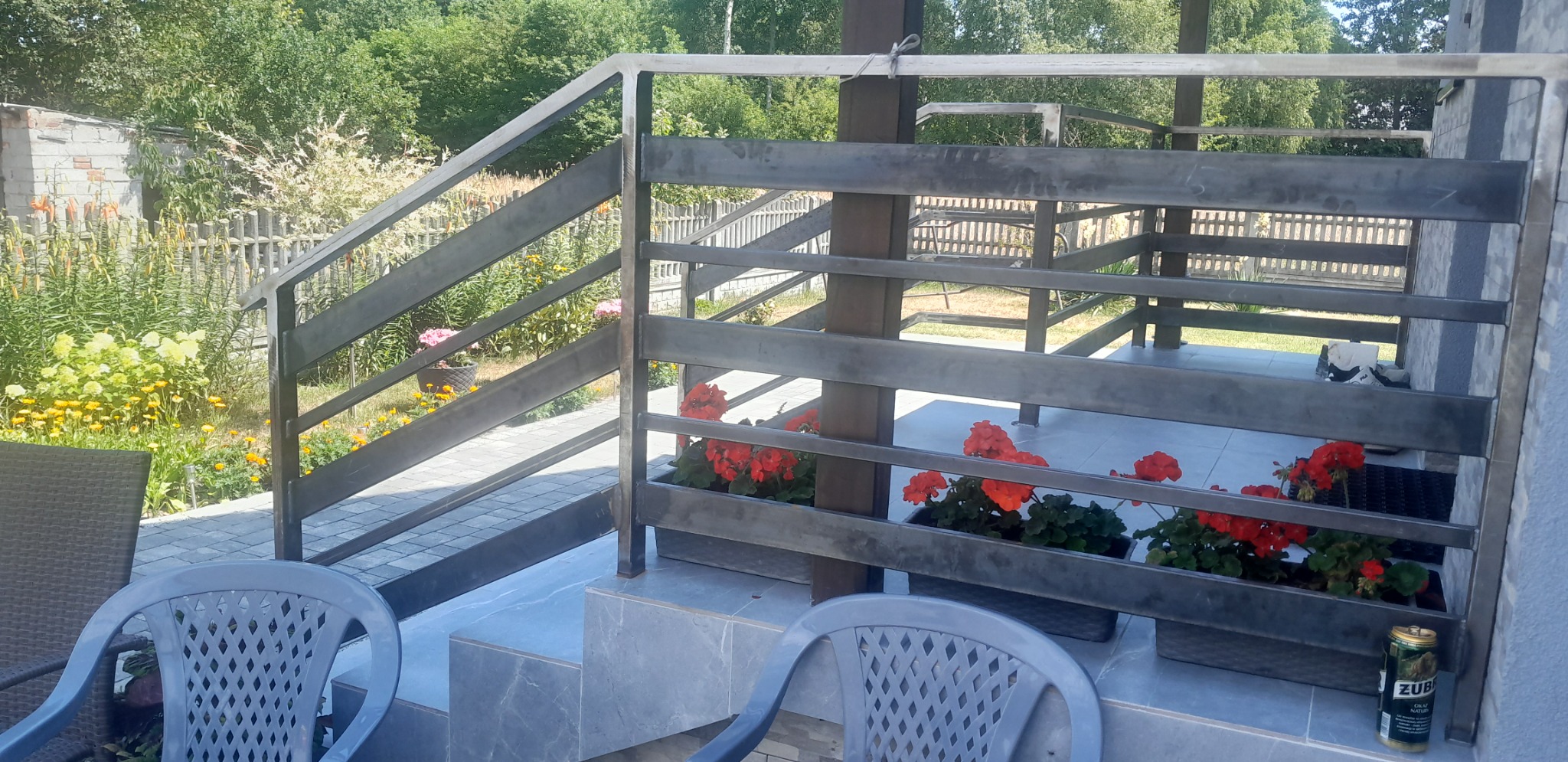 Metalowa balustrada z poziomych profili, donice z czerwonymi kwiatami, widok na ogród z drewnianym płotem, dwa plastikowe krzesła i puszka piwa na posadzce.
