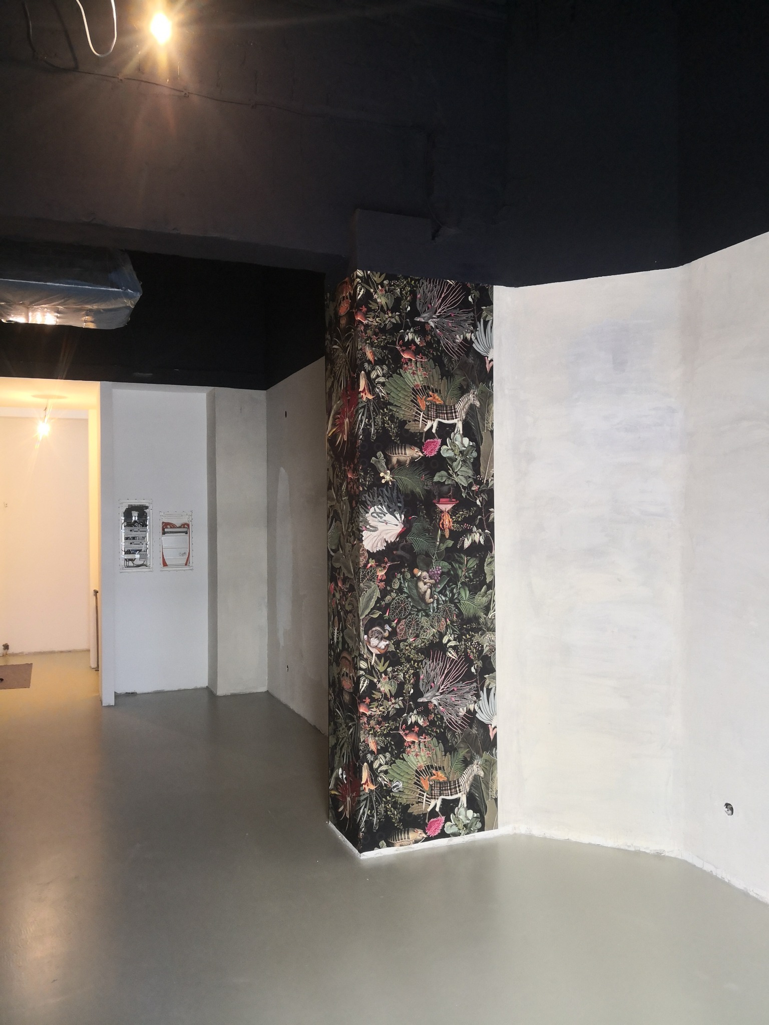 Wnętrze w trakcie prac wykończeniowych: kolumna obłożona tapetą z motywem dżungli, surowa ściana pomalowana na szaro, szara podłoga, czarny sufit, widoczne instalacje elektryczne.