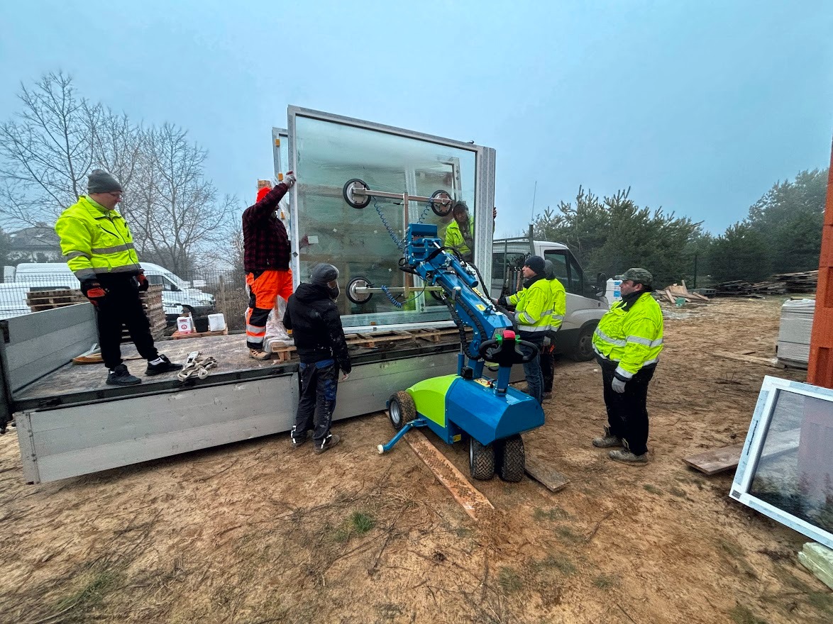 Pracownicy w odblaskowych kurtkach transportują duże okno za pomocą specjalistycznego wózka z przyssawkami na pace samochodu dostawczego.