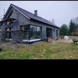 Dom jednorodzinny w trakcie ocieplania, widoczne rusztowania, szara elewacja z dużymi oknami, teren wokół domu częściowo zagospodarowany.