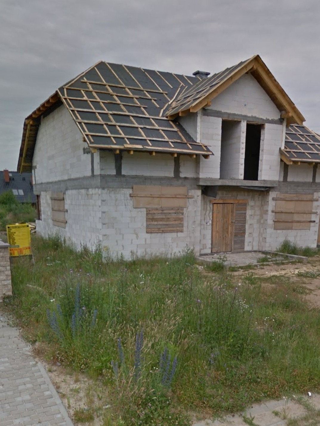 Niedokończony dom z białych bloczków, z częściowo położonym dachem z czarnej papy na drewnianej konstrukcji; okna i drzwi zabite deskami, wysoka trawa przed budynkiem.