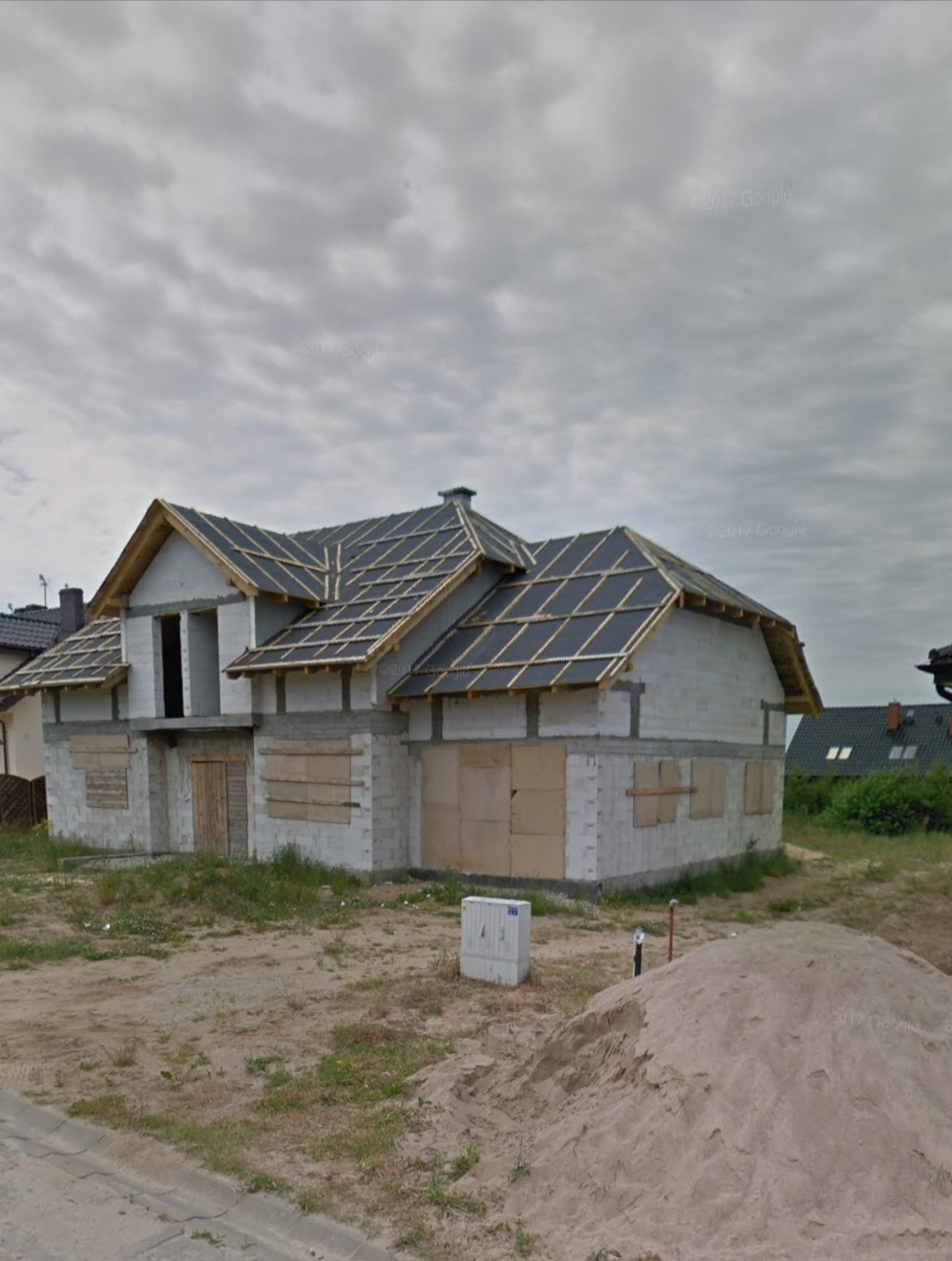 Niedokończony budynek z białych bloczków, z deskami zabitymi oknami i wstępnie pokrytym dachem, widoczny stos piasku na pierwszym planie.