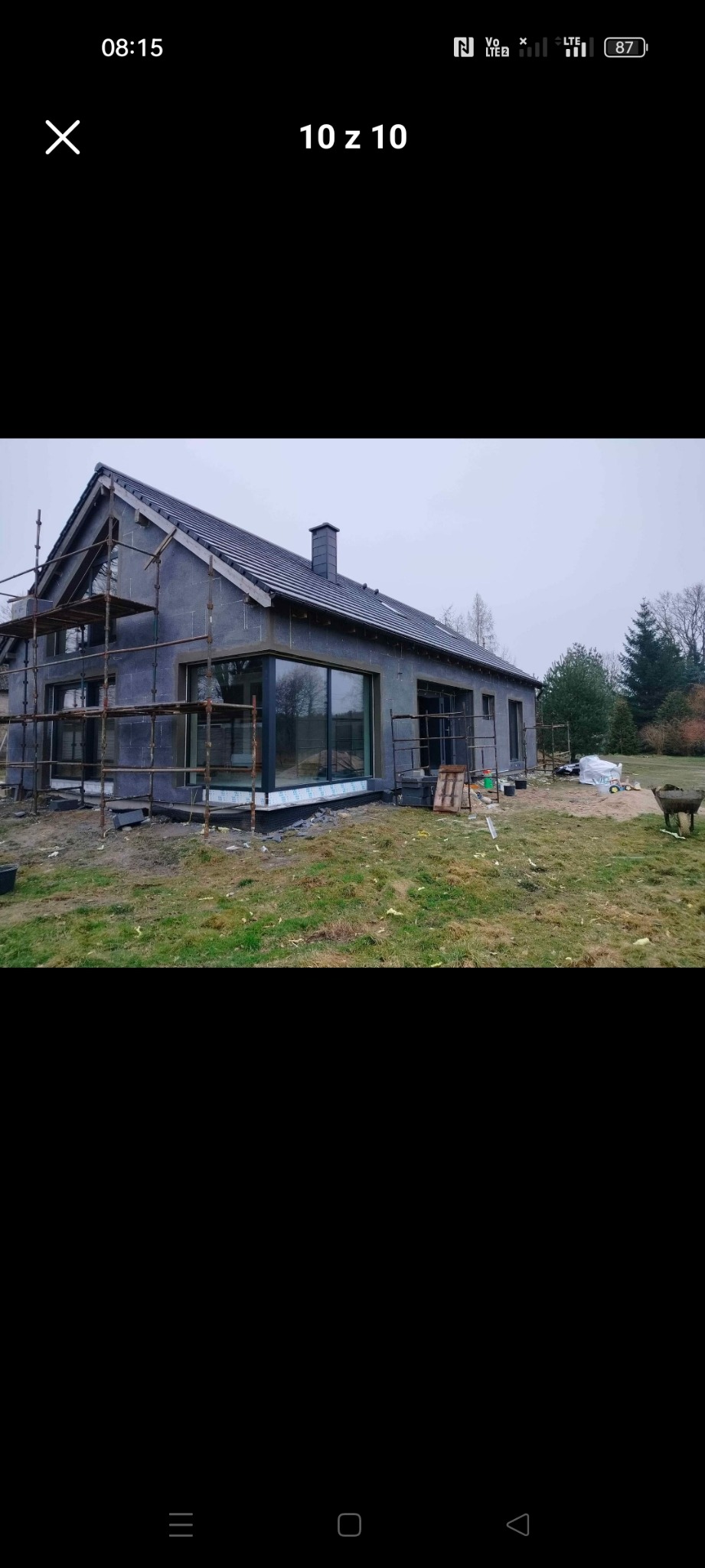 Dom jednorodzinny w trakcie ocieplania, widoczne rusztowania, szara elewacja z dużymi oknami, teren wokół domu częściowo zagospodarowany.