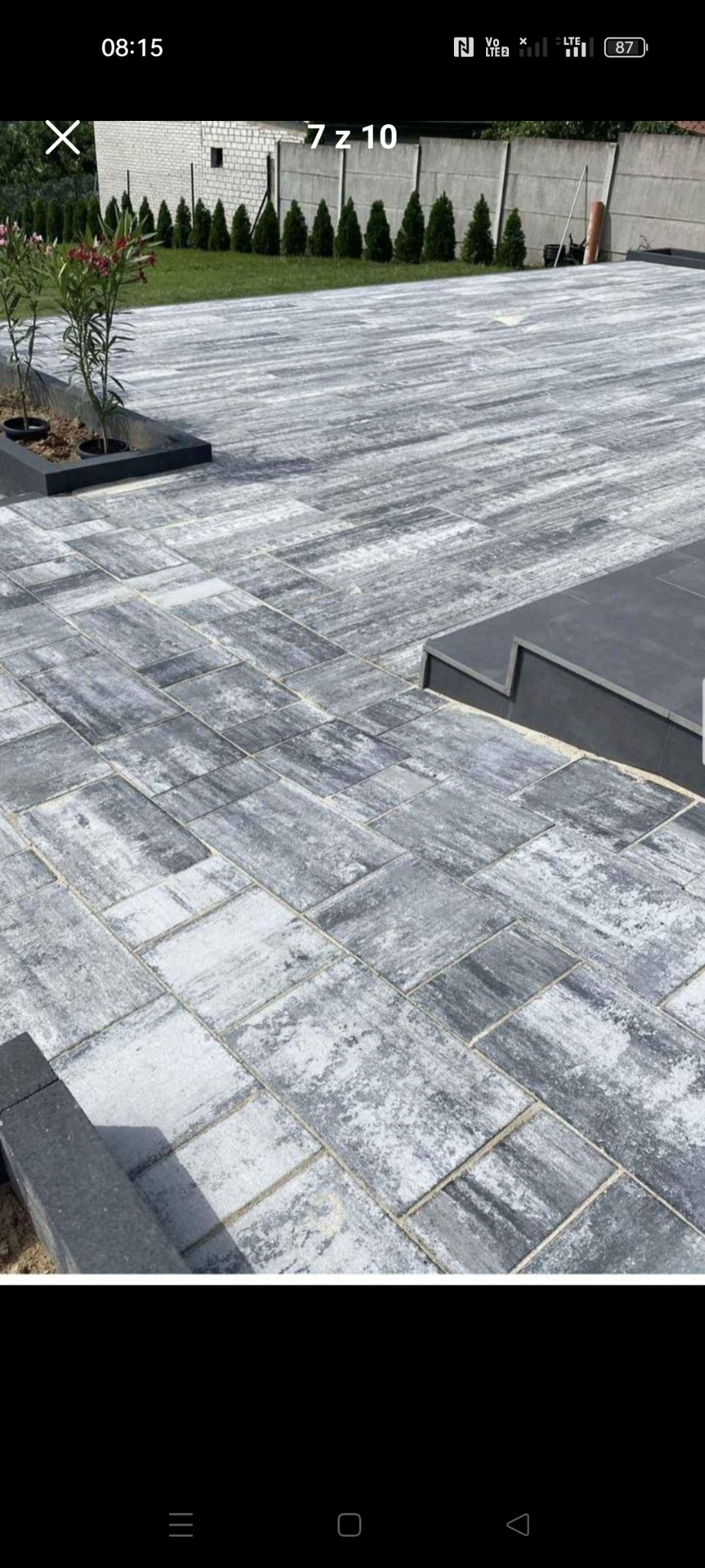 Ułożona szara kostka brukowa w jodełkę z niskimi schodami i roślinami w czarnych donicach, widok z góry na betonowy płot i rząd małych drzewek.