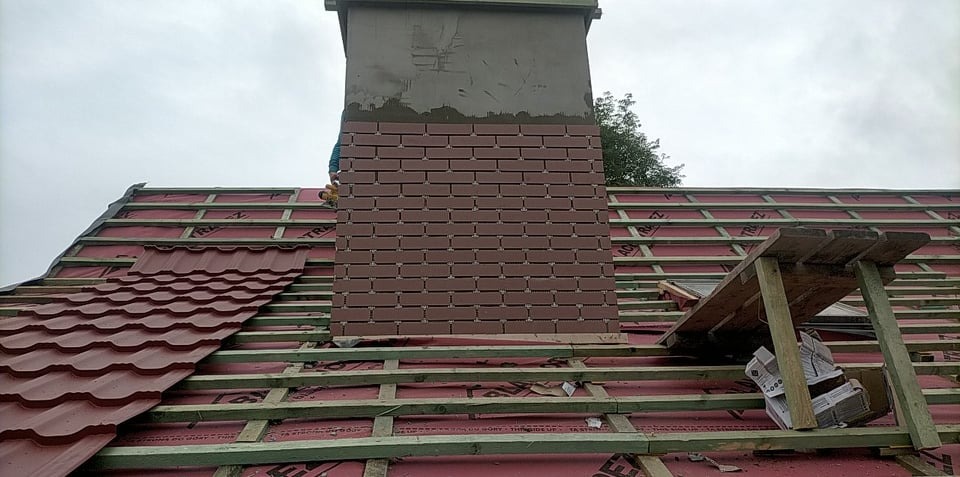 Montaż ceglanej okładziny na kominie dachowym w trakcie budowy dachu z czerwonej blachodachówki, widoczne drewniane łaty i folia izolacyjna.