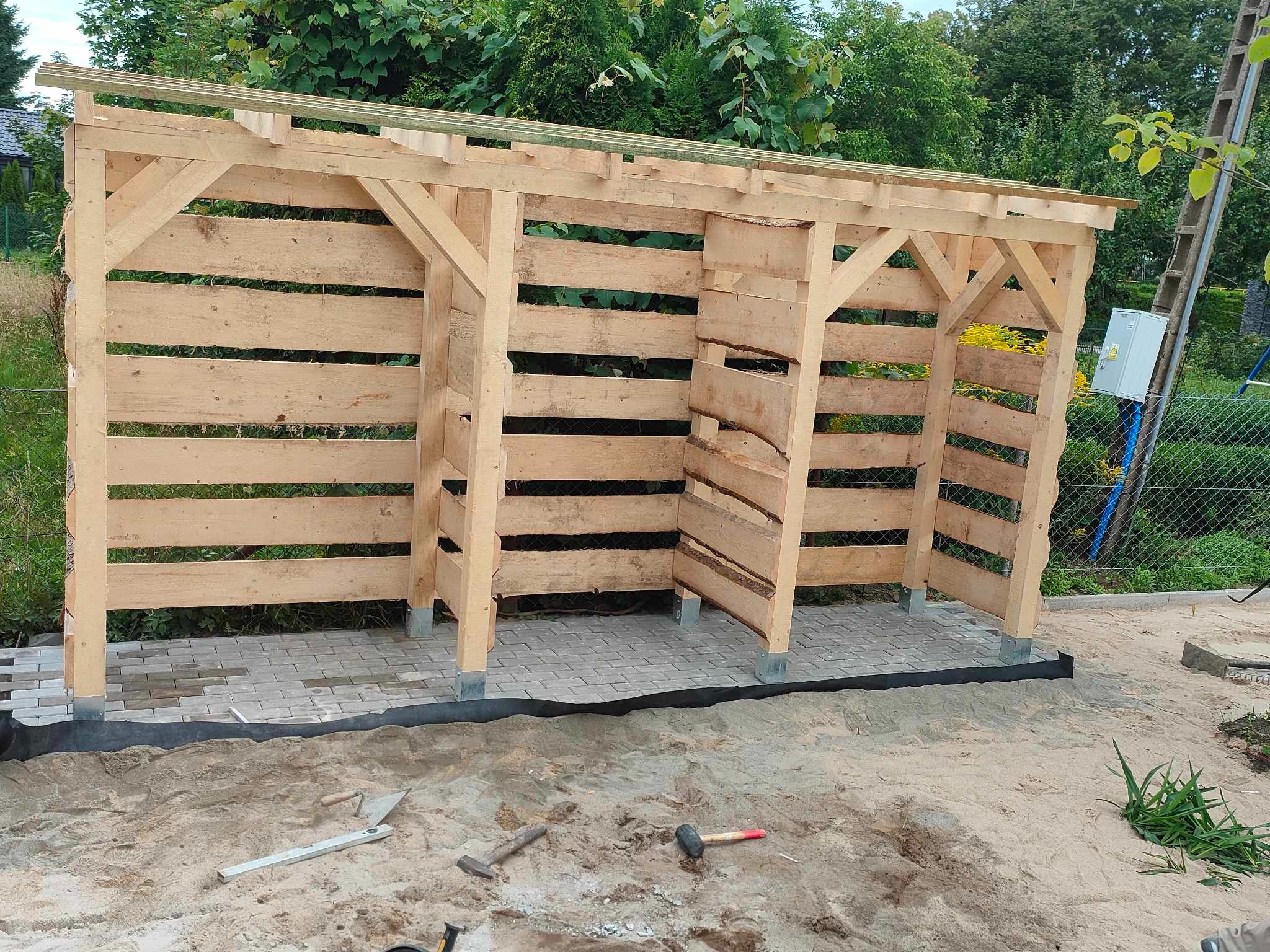 Konstrukcja drewniana w trakcie budowy na podłożu z kostki brukowej, widoczne narzędzia budowlane i piasek.