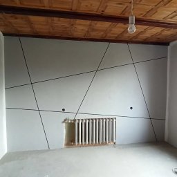 DAMIAN SYKUŁA PROBUD - Wnętrze pokoju z wygładzoną ścianą, na której widoczne są czarne, geometryczne linie dekoracyjne, odsłonięty grzejnik i surowy, drewniany sufit z żarówką.