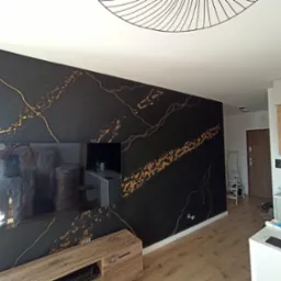 Ściana w salonie z czarną, dekoracyjną strukturą imitującą marmur ze złotymi żyłami, widoczne odbicie malarza w lustrze, nowoczesne oświetlenie sufitowe.