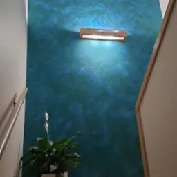 Ściana w odcieniach turkusu i błękitu z efektem chmur, oświetlona drewnianą lampą ścienną, z kwiatem doniczkowym na drewnianych schodach.