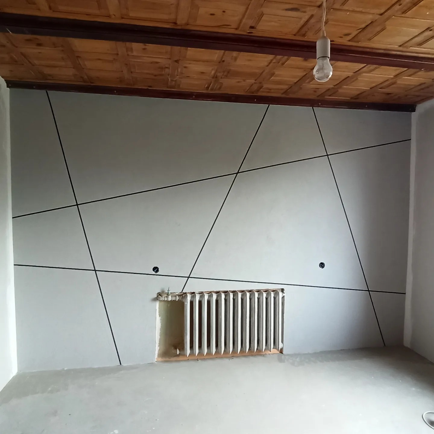 Wnętrze pokoju z wygładzoną ścianą, na której widoczne są czarne, geometryczne linie dekoracyjne, odsłonięty grzejnik i surowy, drewniany sufit z żarówką.