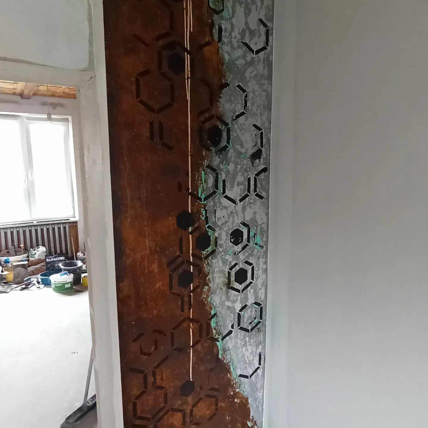 Dekoracyjna ściana z efektem rdzy i wzorem heksagonów w trakcie realizacji, widoczne narzędzia malarskie w tle.