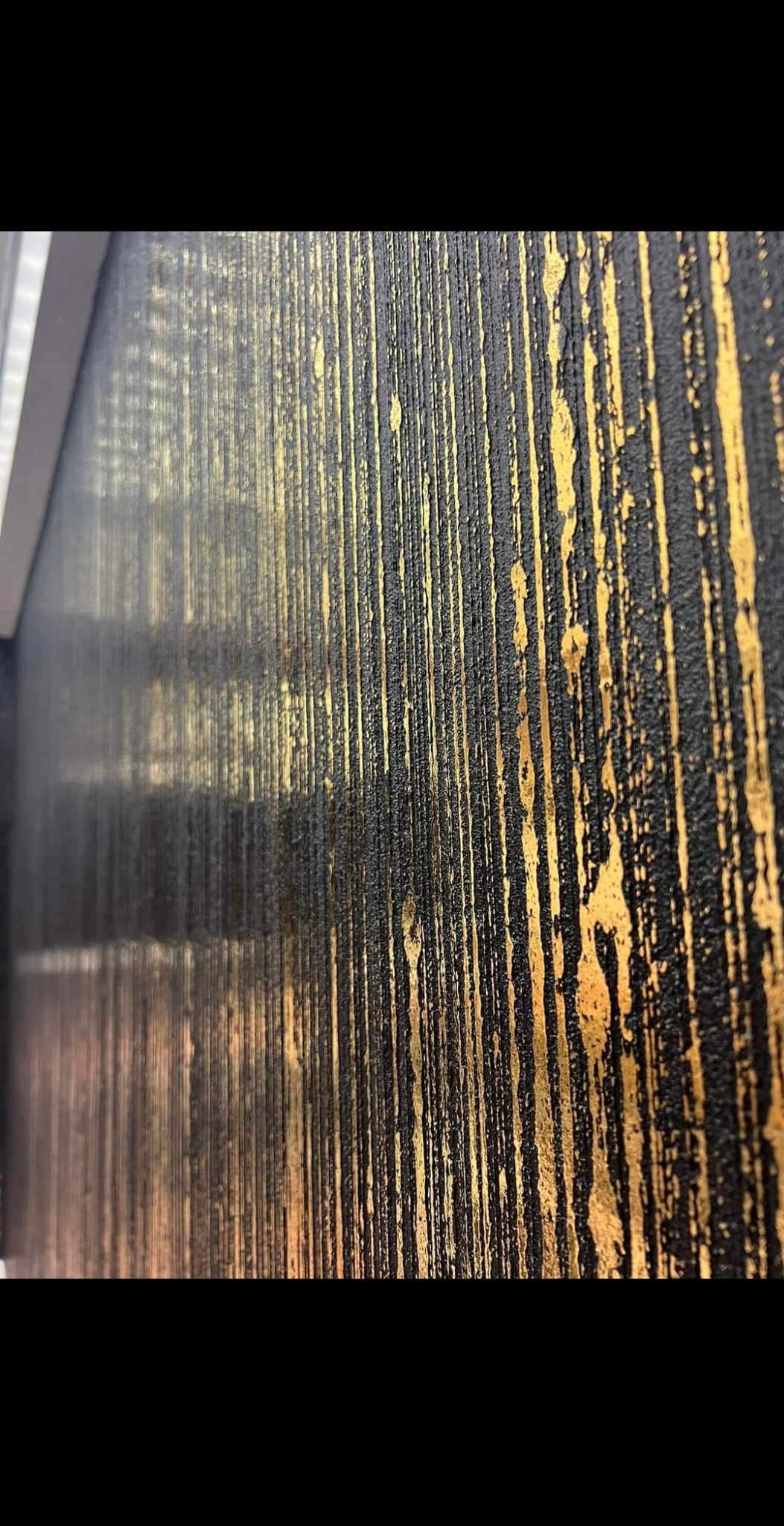 Częściowy widok ściany z dekoracyjną fakturą w kolorze czarnym ze złotymi pionowymi smugami, wykonaną techniką malarską imitującą metaliczną strukturę.