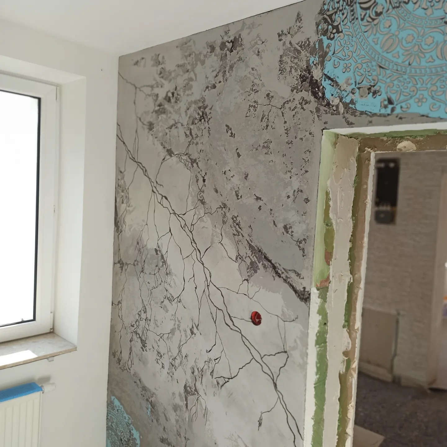 Ściana z dekoracyjną fakturą imitującą marmur z motywem drzewa, fragment nieukończonego wzoru z motywem mandali w rogu, widoczne surowe obramowanie drzwi.