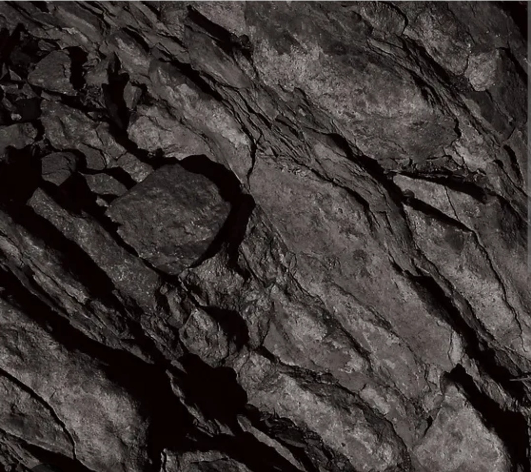 Abstrakcyjna tekstura ciemnoszarej, łupkowej skały z wyraźnymi pęknięciami i warstwami, oświetlona z góry, tworząca mocne kontrasty światła i cienia.