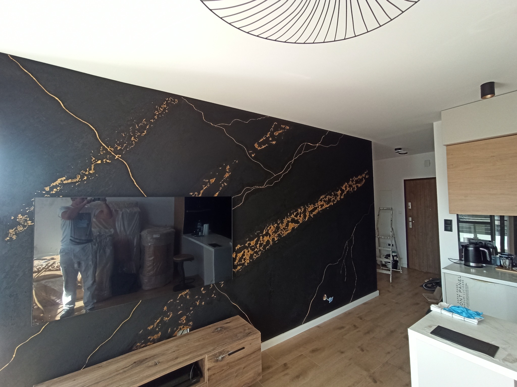 Ściana w salonie z czarną, dekoracyjną strukturą imitującą marmur ze złotymi żyłami, widoczne odbicie malarza w lustrze, nowoczesne oświetlenie sufitowe.