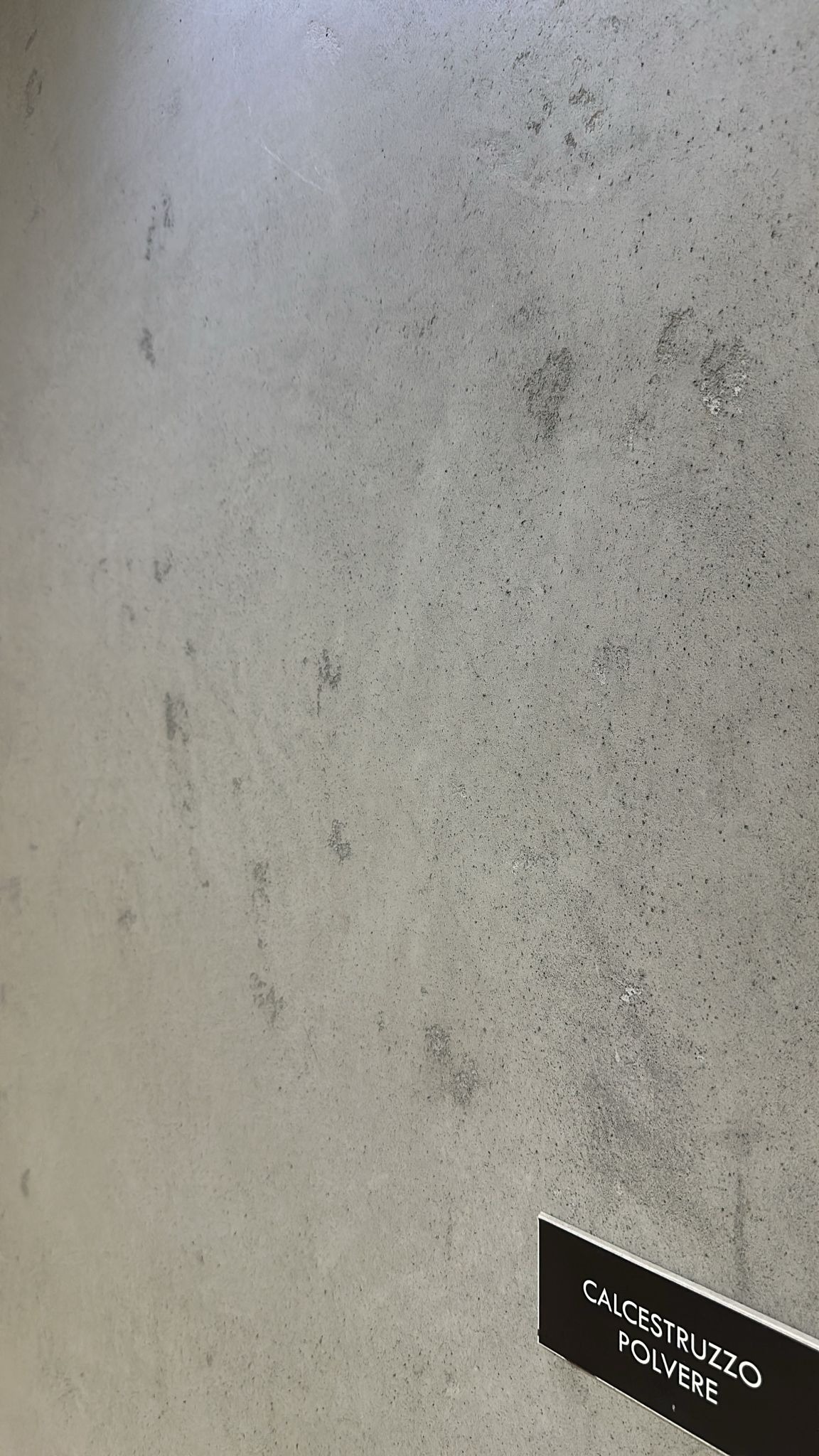 Szara ściana z efektem betonu architektonicznego, z widoczną fakturą i nieregularnymi plamami, oświetlona z góry, z dołączoną etykietą z napisem 'CALCESTRUZZO POLVERE'.