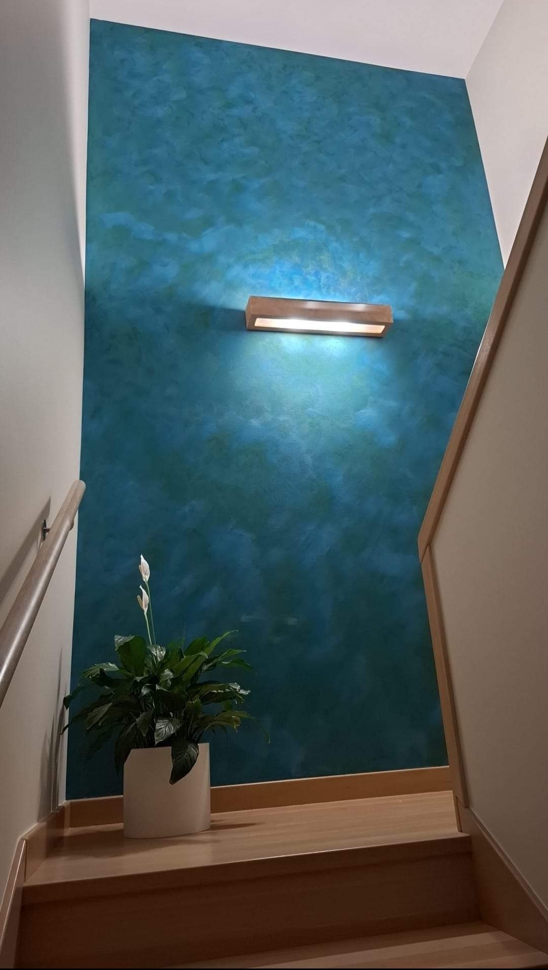 Ściana w odcieniach turkusu i błękitu z efektem chmur, oświetlona drewnianą lampą ścienną, z kwiatem doniczkowym na drewnianych schodach.