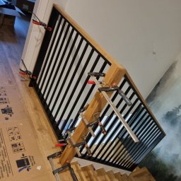 Rafał Lewandowski - Montaż balustrady z czarnymi prętami i drewnianą ramą, mocowanej za pomocą ścisków stolarskich, widoczne schody i tapeta z motywem leśnym w tle.