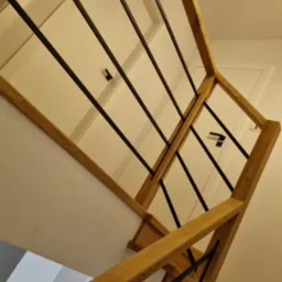 Nowoczesna balustrada schodowa z jasnego drewna i czarnych, pionowych prętów, widziana z dołu, z fragmentem białych drzwi w tle.