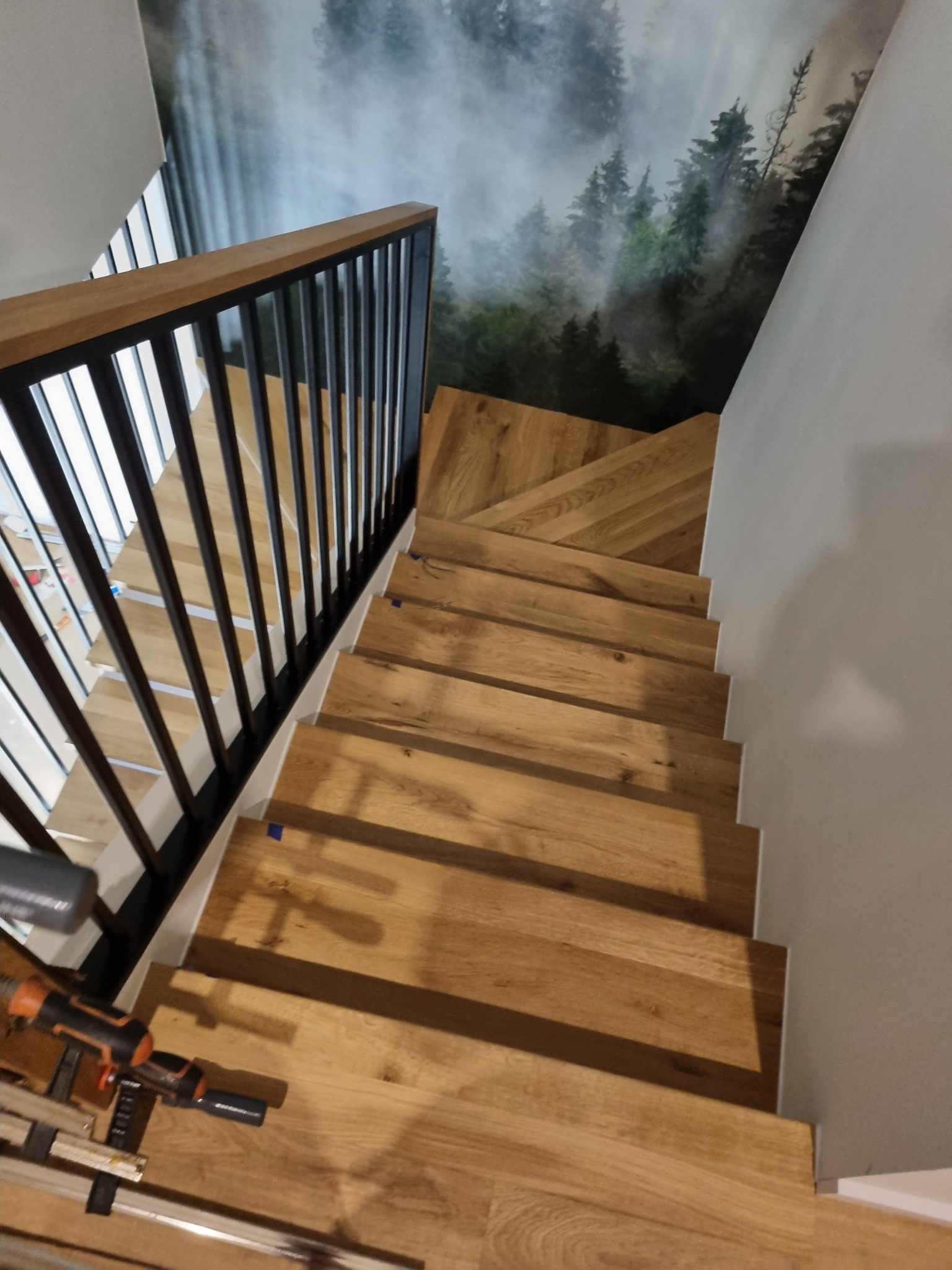 Widok z góry na drewniane schody z czarną metalową balustradą, w tle fototapeta z motywem leśnym, narzędzia do montażu na pierwszym planie.