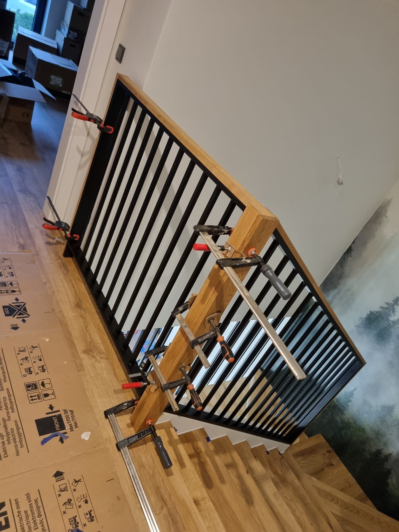 Montaż balustrady z czarnymi prętami i drewnianą ramą, mocowanej za pomocą ścisków stolarskich, widoczne schody i tapeta z motywem leśnym w tle.