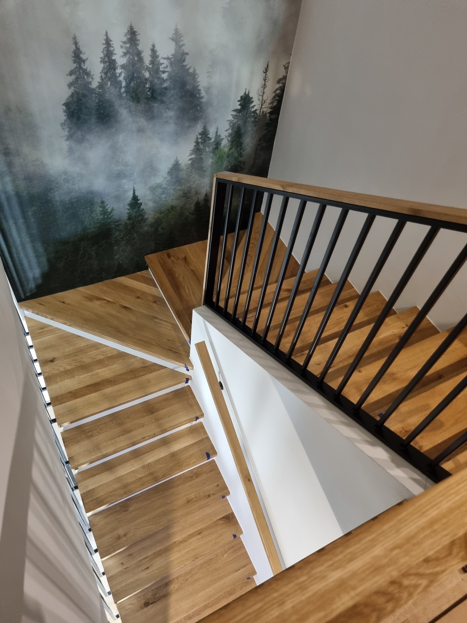 Nowoczesne schody drewniane z czarną metalową balustradą i fototapetą z motywem leśnym na ścianie.