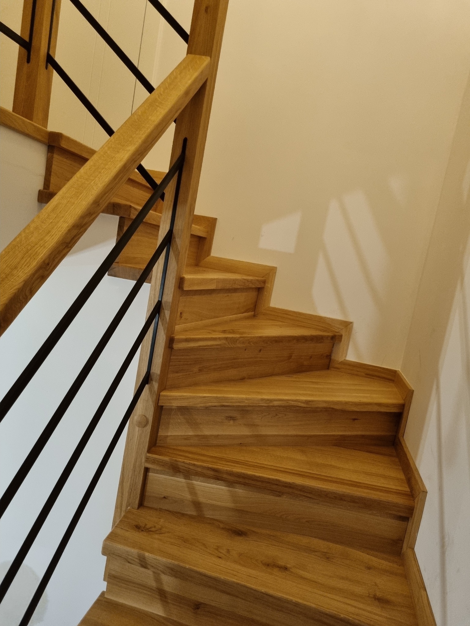 Kątowe ujęcie dębowych schodów z czarną, metalową balustradą, oświetlonych ciepłym światłem, z widoczną strukturą drewna.