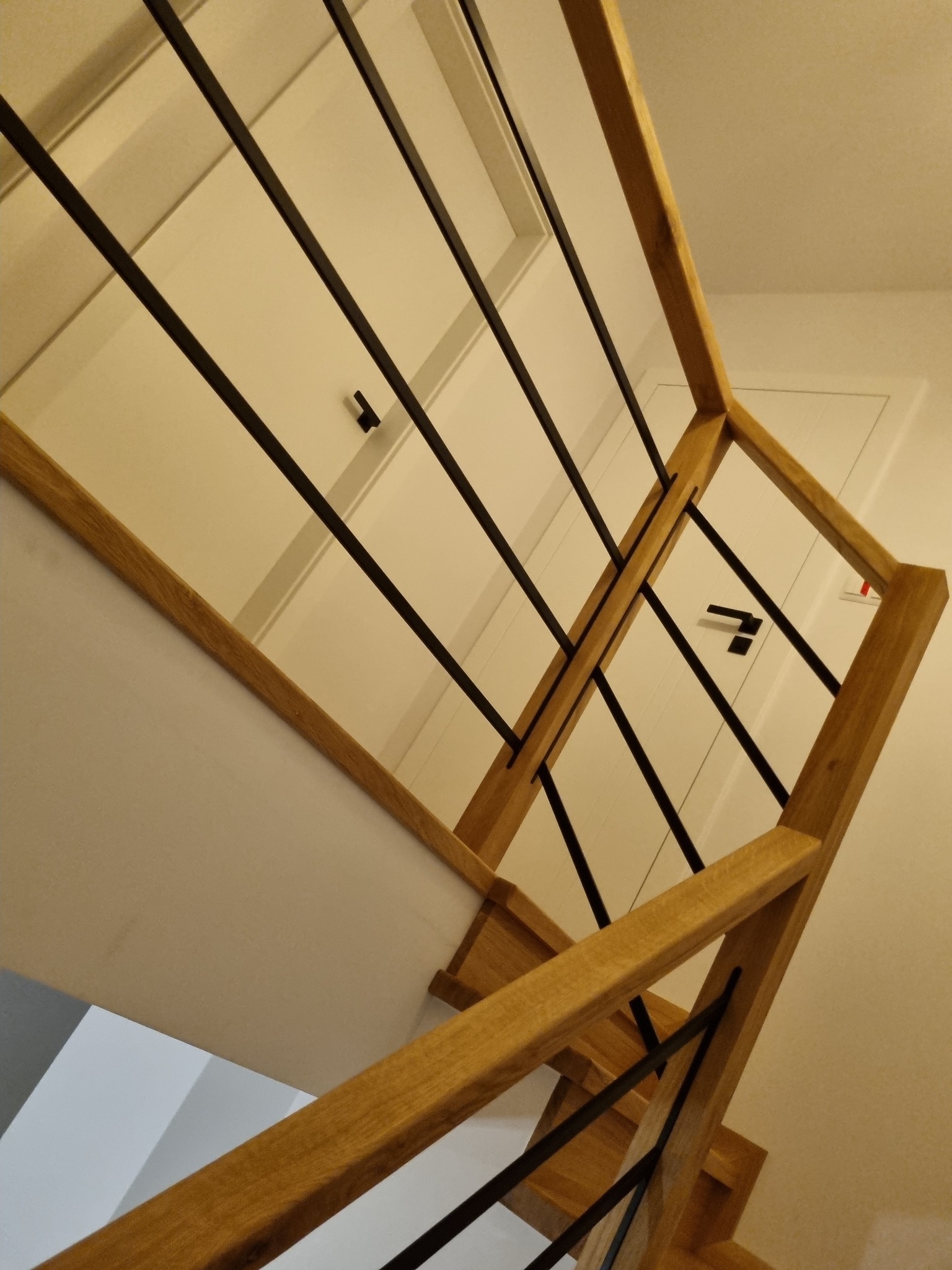 Nowoczesna balustrada schodowa z jasnego drewna i czarnych, pionowych prętów, widziana z dołu, z fragmentem białych drzwi w tle.