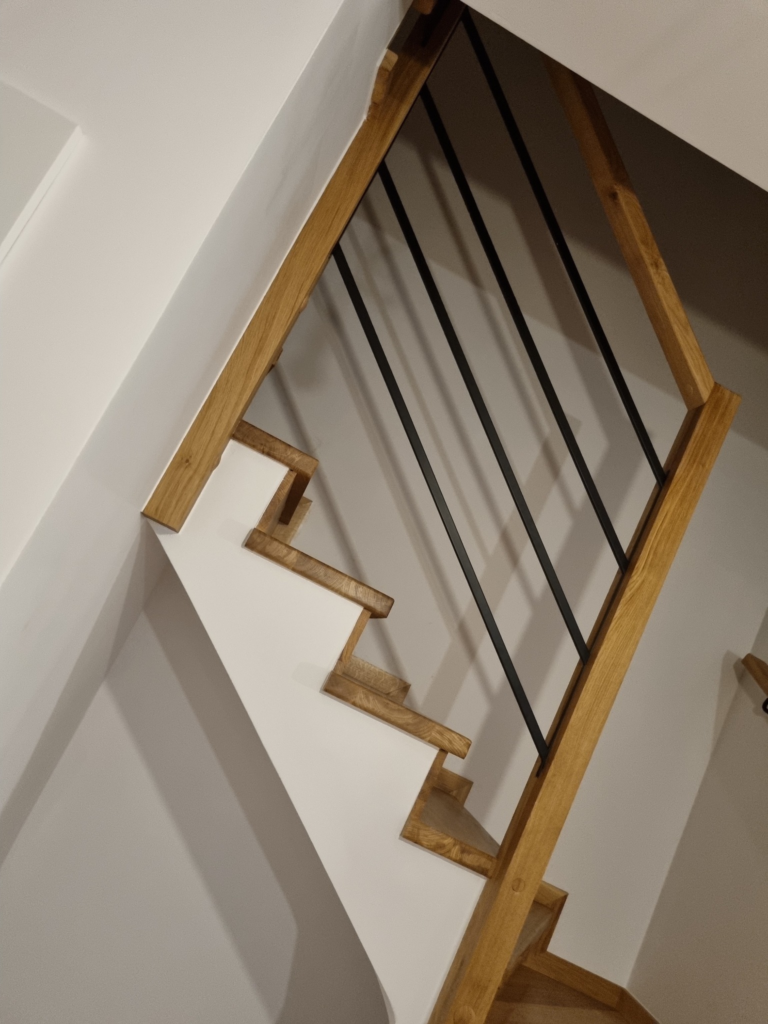 Fragment nowoczesnych schodów z drewnianymi stopniami, białymi podstopnicami i czarną, metalową balustradą, widziany z dołu pod kątem.
